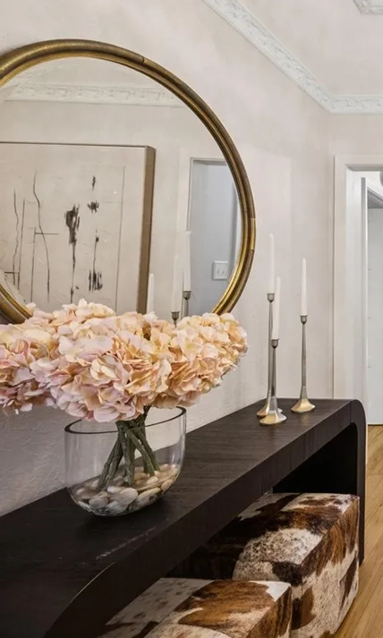 Entryway styling featuring a dark console table with peach hydrangeas under a round gold mirror and two rawhide upholstered ottomans