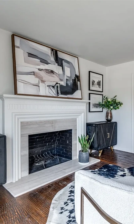 Close-up of a white wooden fireplace mantel with large abstract art above it and a dark wood accent cabinet to the side