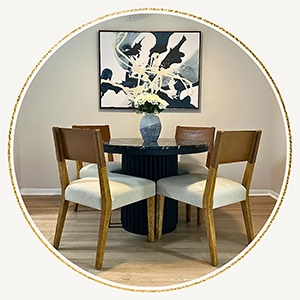 Circular vignette showing a round black fluted dining table with four modern wood and fabric chairs under an abstract art piece