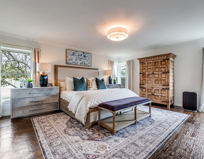 Staged primary bedroom featuring an upholstered bed, a purple velvet bench, a rustic wood armoire, and a patterned rug