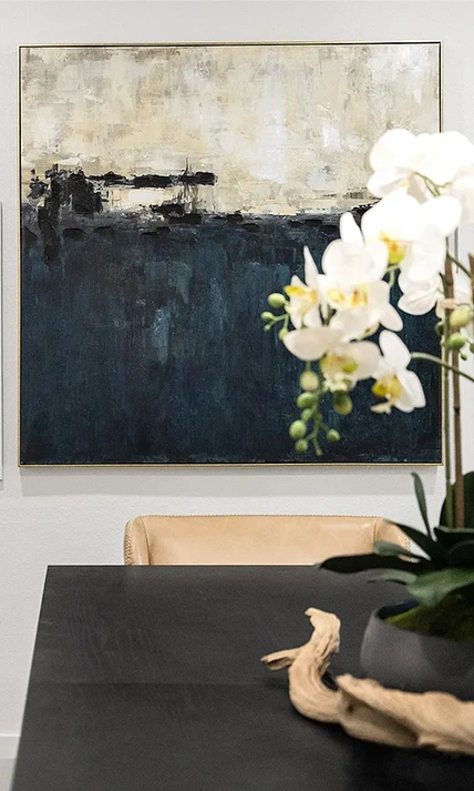 A close-up of a large-scale abstract painting with deep navy, cream, and black tones hanging behind a tan leather chair and a white orchid centerpiece.
