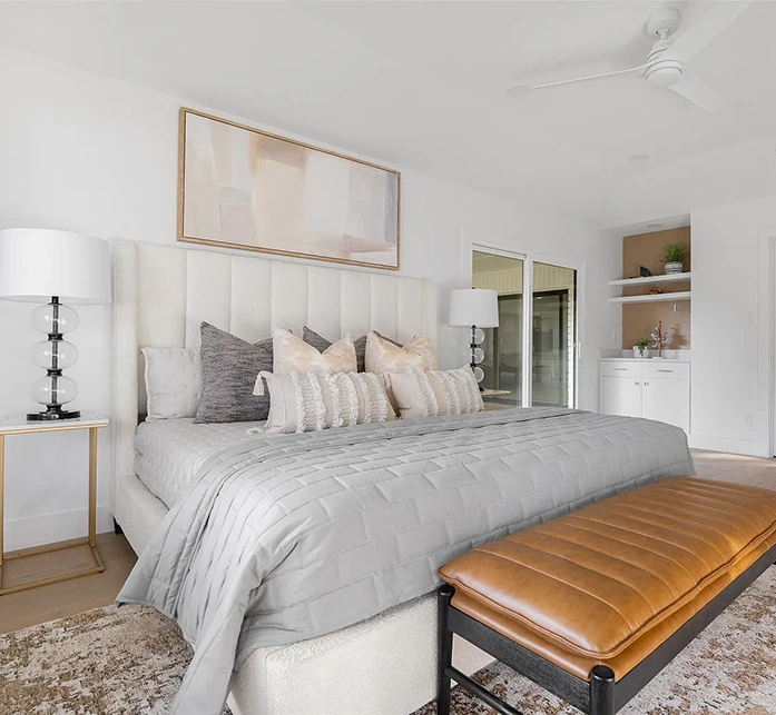 A luxurious bedroom featuring a tall, cream-colored channel-tufted headboard and a bed dressed in layered grey and white linens. A cognac leather bench sits at the foot of the bed on a patterned rug, with a large neutral abstract painting hanging on the wall above.