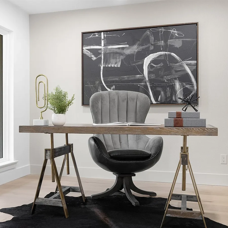 A modern home office setup with a wood-top desk on metal sawhorses. A grey velvet tufted swivel chair sits on a black cowhide rug. On the wall behind, a large black and white abstract painting adds a bold focal point to the bright space.
