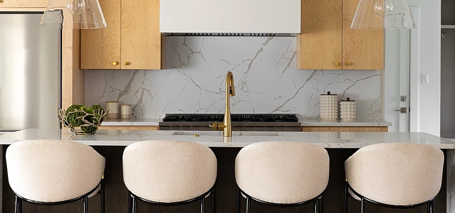 A close-up of a kitchen island with a white marble waterfall edge and a gold faucet. Four white bouclé barstools are tucked under the counter, set against a background of warm light-wood cabinets and a white marble backsplash.