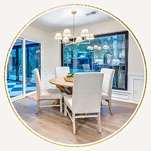 A circular photo of a round wooden dining table with four high-back cream chairs, situated in front of a large window overlooking a lush backyard.