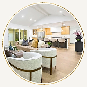 A circular photo showing the flow from white textured armchairs in the foreground to a large tan sofa and a modern kitchen in the background under a vaulted ceiling.