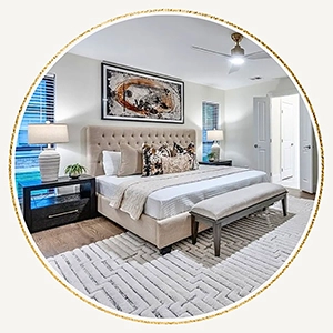 A circular photo of a luxurious bedroom featuring a beige tufted headboard, a grey quilted duvet, a cognac leather bench, and a large abstract painting above the bed. 