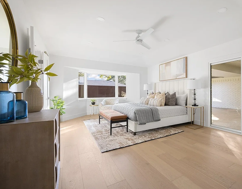 A spacious, white-walled bedroom with a large bay window, featuring a white upholstered bed with layered grey and gold pillows, a cognac leather bench, and a modern white ceiling fan.