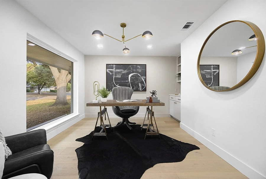 A bright office featuring a rustic wood desk on sawhorses, a grey tufted executive chair, and a black cowhide rug over light wood floors. A large circular gold mirror reflects the room, and a black and white abstract painting hangs on the back wall.