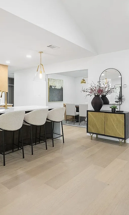 A view from the kitchen island showing white bouclé barstools, wood-toned cabinetry with a white marble backsplash, and a glimpse into a dining room with a large black vase and floral arrangement.