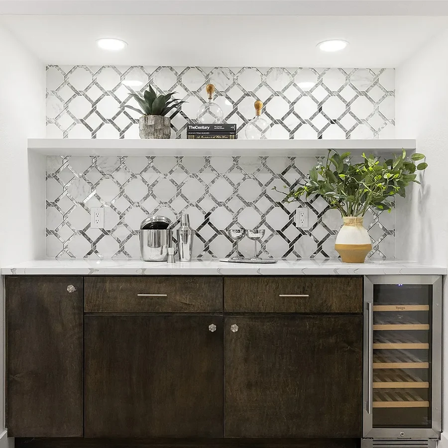 A sophisticated home bar area with dark wood cabinets, a built-in wine fridge, white stone countertops, and a reflective silver geometric tile backsplash adorned with minimalist decor and greenery.