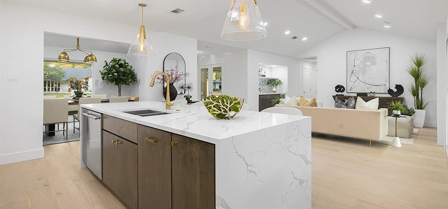 A wide white marble waterfall island with gold fixtures and four white upholstered barstools, set against warm wood upper cabinets and a professional gas range.