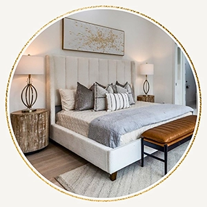 A circular image featuring a professionally staged bedroom with a cream-colored channeled headboard, neutral bedding, a leather bench at the foot of the bed, and round wood nightstands with elegant lamps