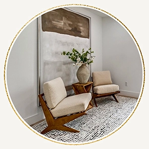 A circular image of a minimalist sitting area with two mid-century modern wooden armchairs, a geometric wood side table, and a large abstract painting on the wall