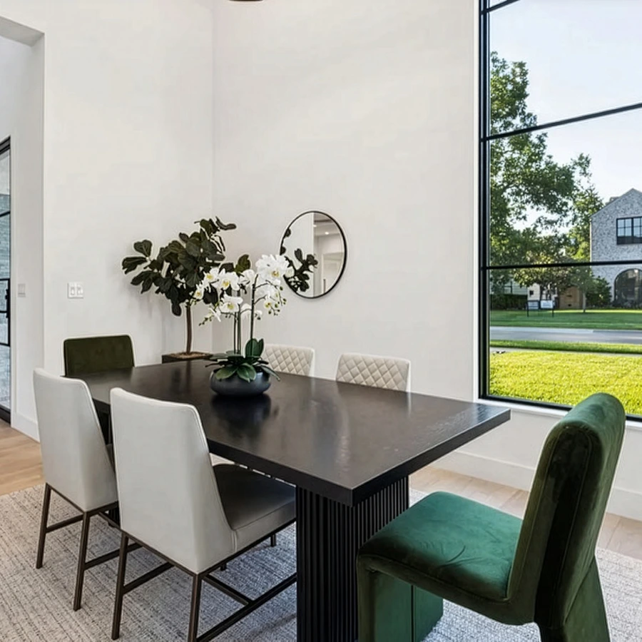 Brightly staged dining room in Prestonwood with a black rectangular dining table, green velvet end chairs, and a large window overlooking a lush green lawn