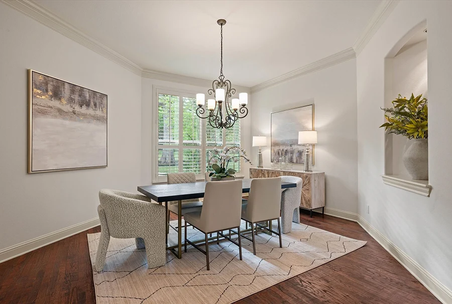 A traditional dining room with dark wood floors, featuring a long black table with six grey upholstered chairs. A wrought iron chandelier hangs above, and a large piece of abstract art is flanked by two white lamps on a sideboard.