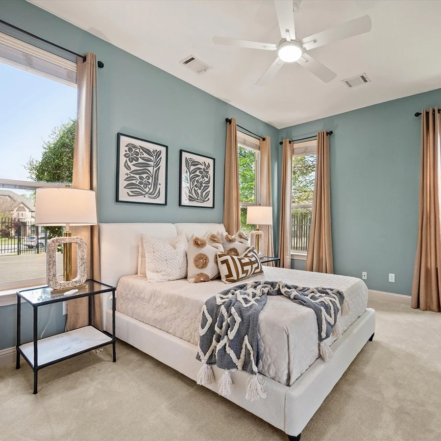 A bright guest bedroom with soft blue walls, featuring a white upholstered bed with patterned brown and white pillows and a blue patterned throw. Large windows with neutral drapes let in abundant natural light.