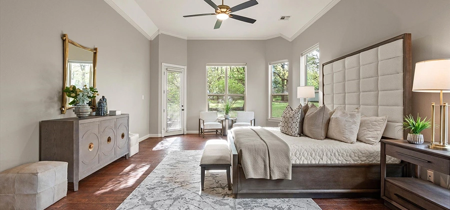 Modern, large primary bedroom with grey upholstered bed with dark wood surrouding the upholster, white and grey bedding, large dresser with mirror and white ottomans.