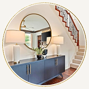 A circular photo of a navy blue console table positioned under a large round mirror near the staircase. Two white textured lamps flank a small green plant and a decorative black and white vase.