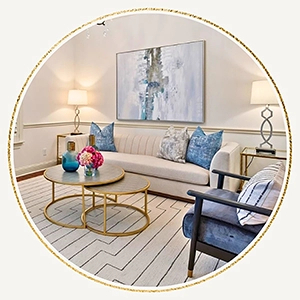 A circular photo showing a close-up of a light-colored sofa with blue patterned pillows, a gold circular coffee table with a floral arrangement, and a blue velvet armchair.