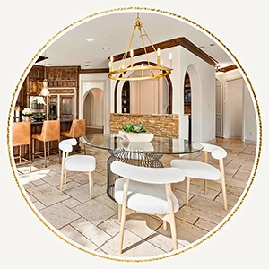 A circular photo centered on a modern glass dining table with a black wire base and white textured chairs, set against the backdrop of the home's stone and wood kitchen architecture.