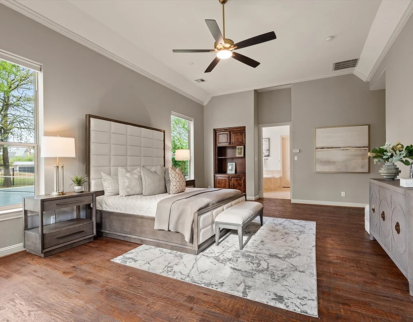 A grand bedroom with vaulted ceilings and a large windows. It features a grey upholstered bed with a tall square-tufted headboard, a white bench at the foot of the bed, a grey wood nightstand, and a matching grey dresser with gold hardware. A large neutral landscape painting hangs on the far wall.
