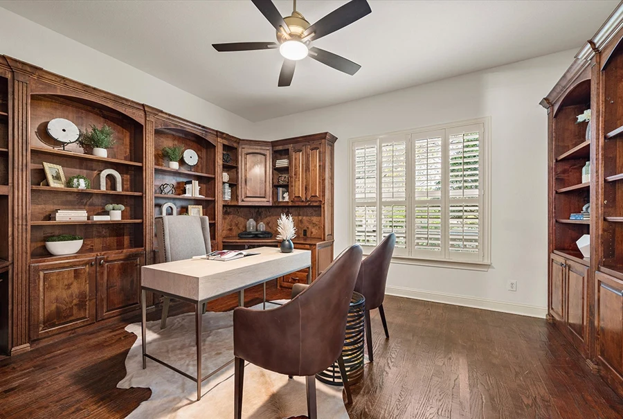 A rich office space featuring floor-to-ceiling dark wood built-in bookshelves and cabinetry. A modern white desk sits on a cowhide rug, paired with a grey upholstered chair and two cognac leather armchairs.