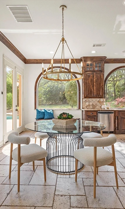 A bright dining area featuring a round glass-top table with a black wire base, surrounded by four white bouclé chairs. The room has large windows, light wood floors, and opens into a kitchen with dark wood cabinetry and a gold ring chandelier.