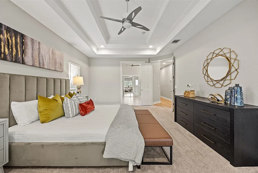Staged bedroom with grey upholstered bed, tan bench, black dresser and large round mirror