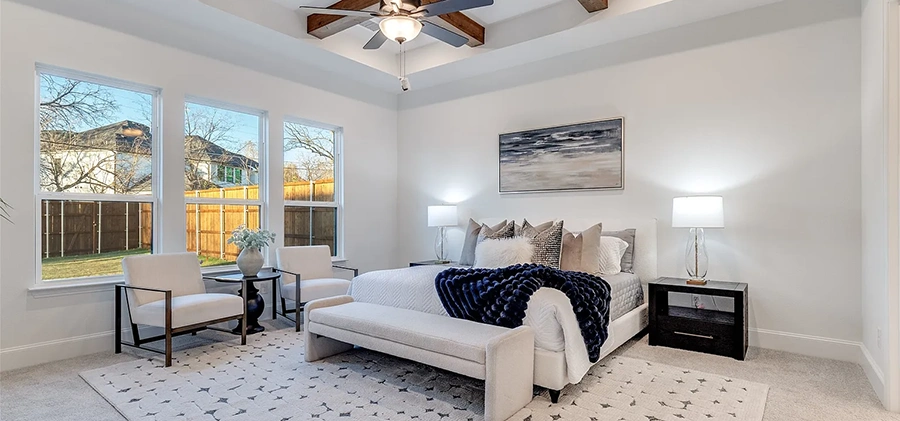 Staged bedroom with white upholstered bed, two white accent chairs, blue and white pillows and artwork