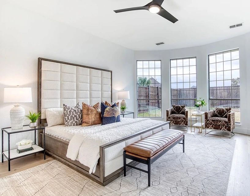 Luxury staged bedroom with a large upholstered bed, plush bedding, leather bench and two modern brown and white chairs