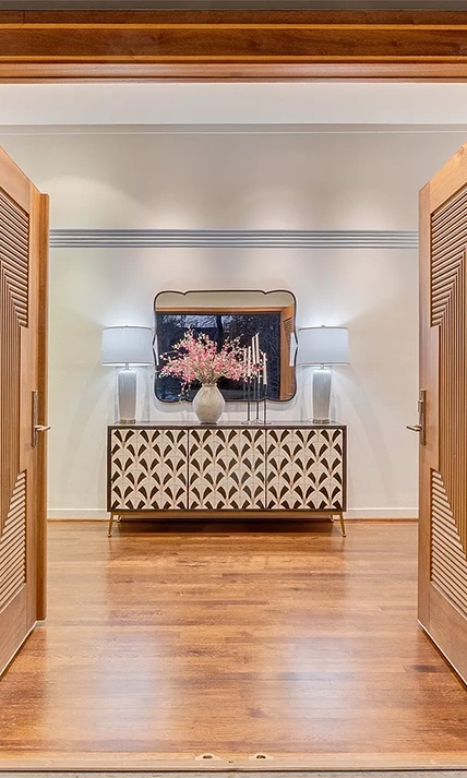 Entryway staged with a large white a light wood console table, a large mirror, two white lamps and a white vase with pink flowers
