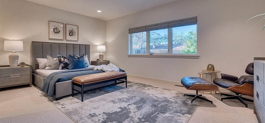 Bedroom staged with a large grey upholstered bed, light tan leather bench, grey nightstands and blue and white accents