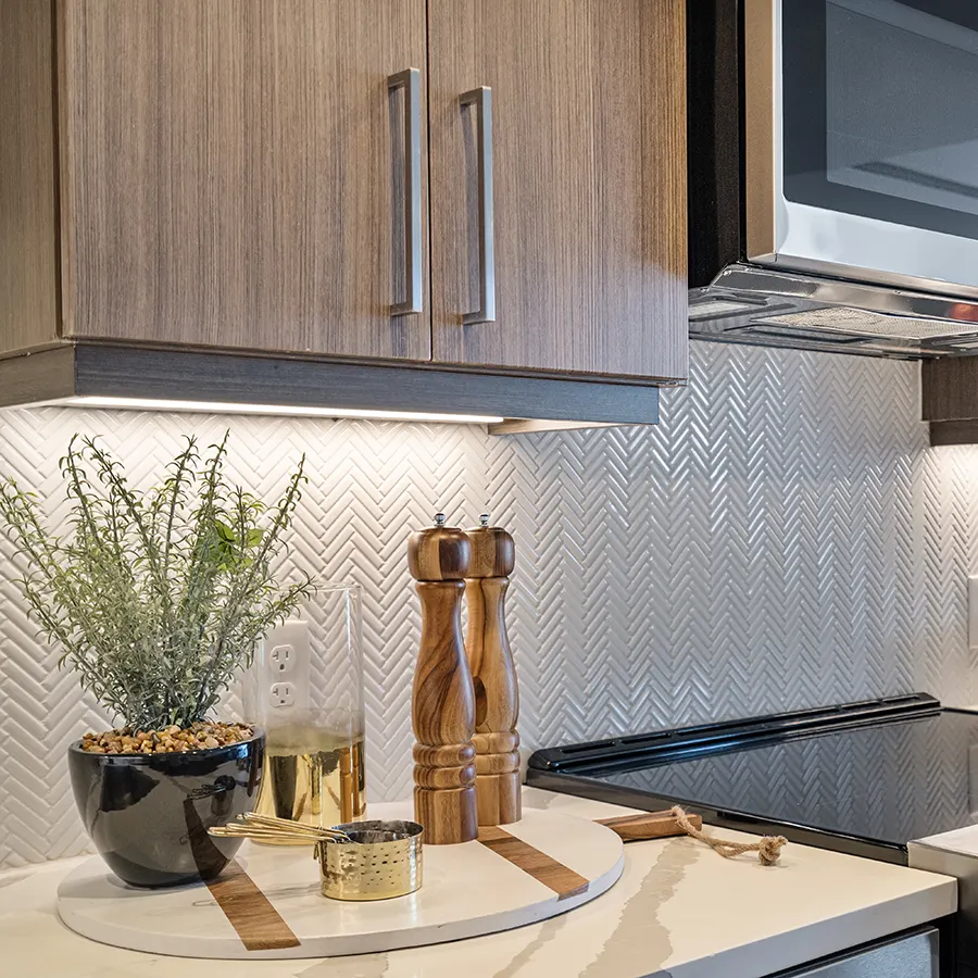 Detailed view of a modern kitchen counter with a white herringbone backsplash, wooden pepper mills, and a small potted plant under cabinet lighting