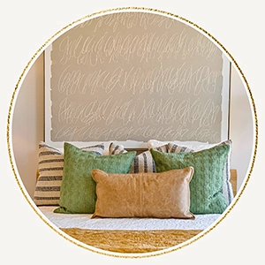 
            Circular inset image of a bed with layered pillows in neutral, green, and tan tones, set against a large piece of abstract script wall art