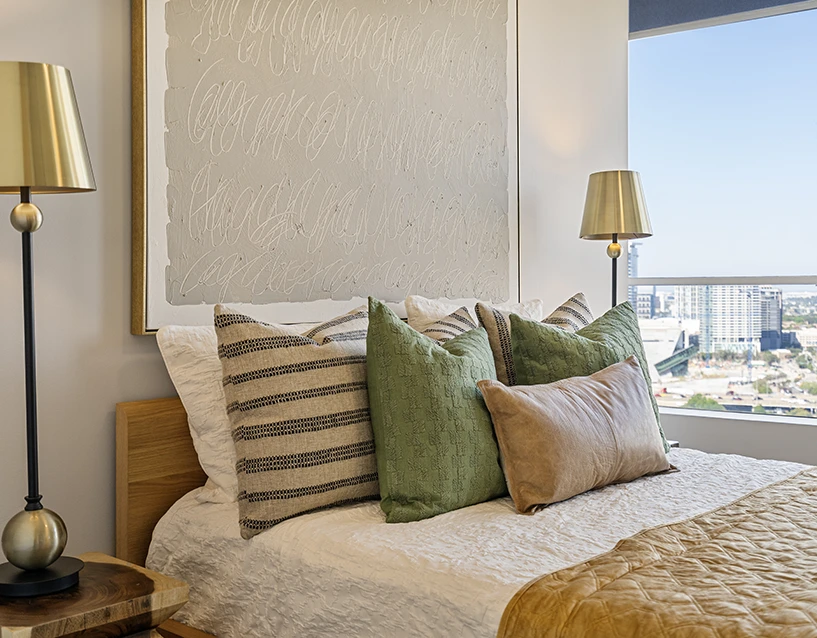 Modern bedroom staging with a wooden bed frame, layered neutral and green pillows, and brass bedside lamps overlooking a Dallas urban view