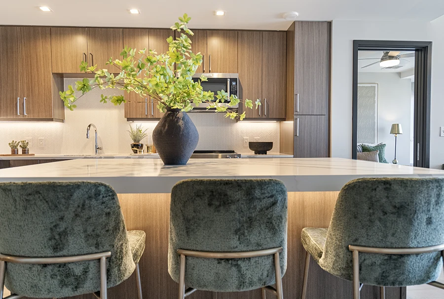 A white marble breakfast bar with three green velvet stools positioned in front of a modern kitchen with wood-toned cabinetry and under-mount lighting