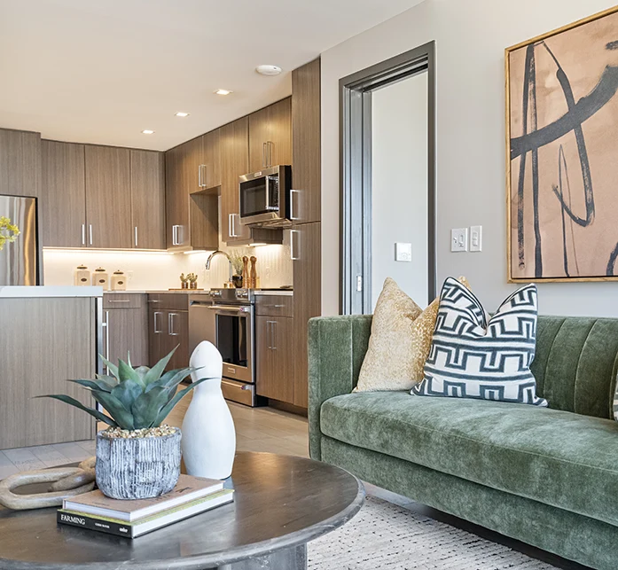 View from a staged living area showing a green velvet sofa and contemporary coffee table with a glimpse into a modern kitchen with wood cabinetry