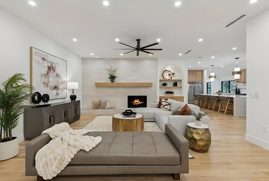 Staged wide-open living room highlighting a tufted grey leather daybed with a plush cream throw blanket, a light grey corner sectional, a round wooden coffee table, and an open transition into the kitchen