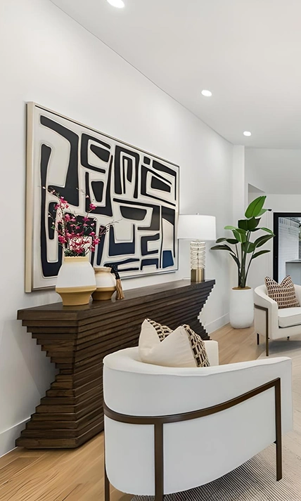 Close-up of a staged hallway featuring an architectural tiered dark wood console table, a large black and white abstract canvas painting, a glass table lamp, and a white modern armchair