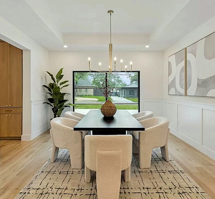 Staged dining room with a long dark wood table, six textured cream chairs, a minimalist gold candle-style chandelier, and a large window looking out to the front yard