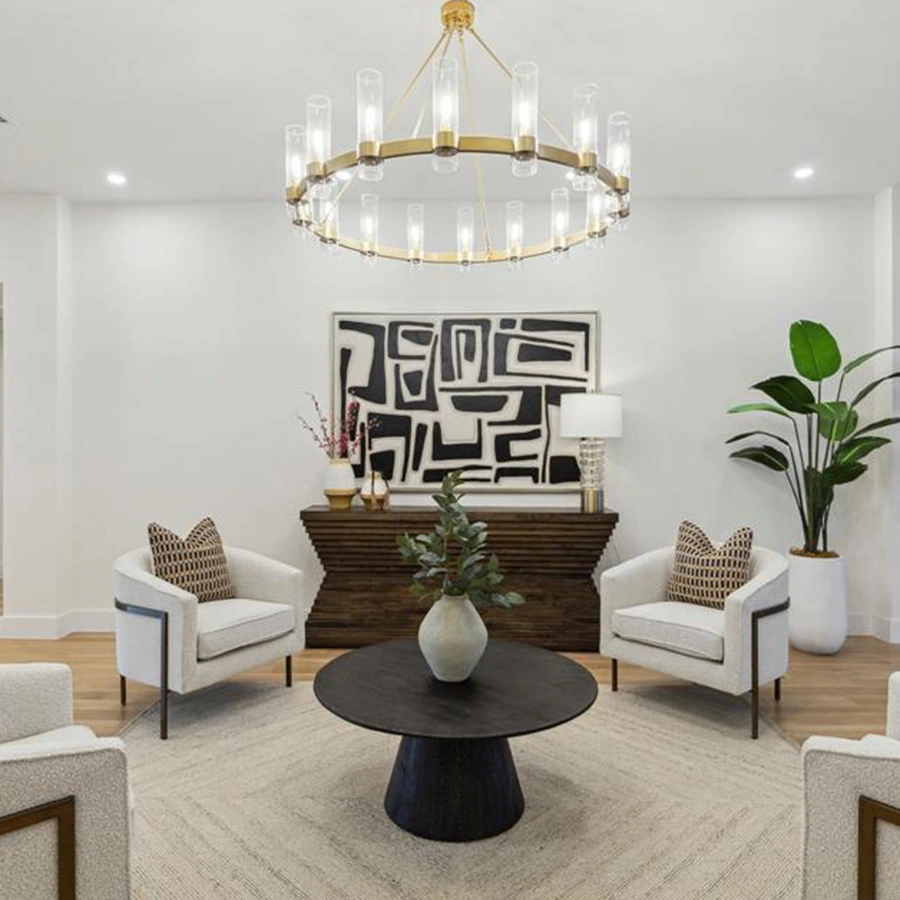 Staged living area detail showcasing a low-profile round black coffee table on a neutral herringbone area rug, flanked by two modern white club chairs beneath a large gold multi-light ring chandelier