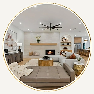 Circular image showcasing a staged living room with a stone fireplace, a grey leather daybed, a light grey sectional, and an open floor plan looking toward the kitchen