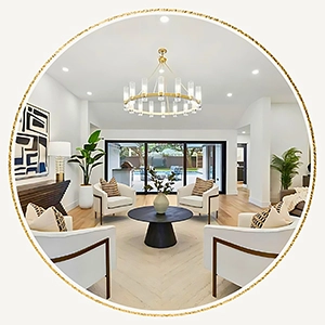 Circular image showcasing a staged open-concept living room with white accent chairs surrounding a black coffee table, a gold ring chandelier, and large sliding glass doors leading outside