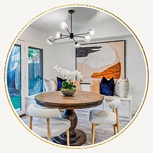 Circular image showcasing a staged breakfast nook with a round wood pedestal table, cream textured chairs, a built-in window bench with blue velvet pillows, and a modern black sputnik chandelier