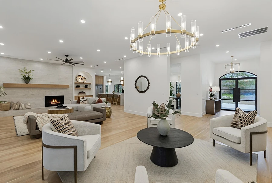 Staged living room highlighting a black pedestal coffee table on a neutral textured rug, cream swivel chairs, a decorative dark wood circular mirror, and an open layout leading to the kitchen