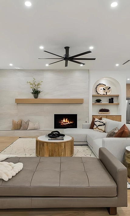 Staged living area featuring a sleek floor-to-ceiling stone fireplace with a light oak mantel, an arched built-in shelving alcove, a light grey sectional, and a grey leather tufted daybed
