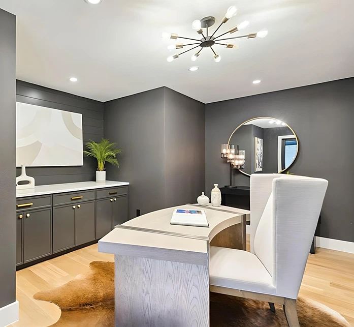 Staged contemporary home office with deep charcoal grey walls, a light wood executive desk, a white high-back wingback chair, and a brown-and-white cowhide area rug