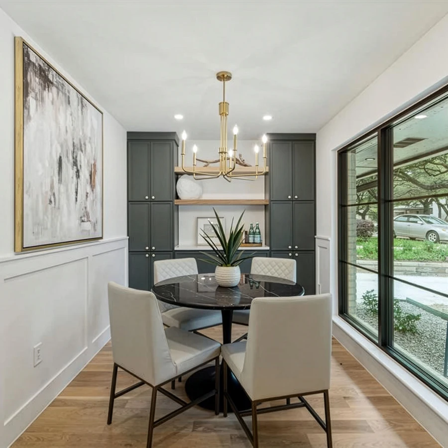 Staged dining space featuring a round black marble-top table, four cream upholstered dining chairs with dark legs, a modern gold geometric chandelier, and floor-to-ceiling charcoal gray built-in cabinets