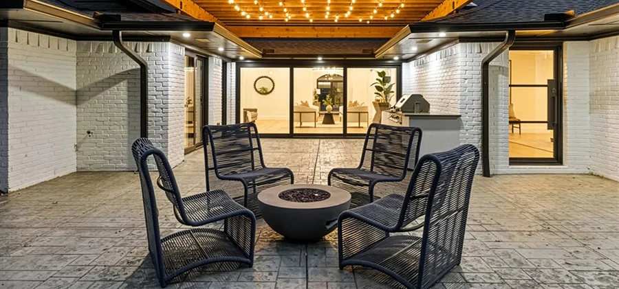 Staged outdoor concrete patio showcasing a round grey fire bowl, four navy blue woven strap lounge chairs, white painted brick exterior walls, and warm overhead string lighting